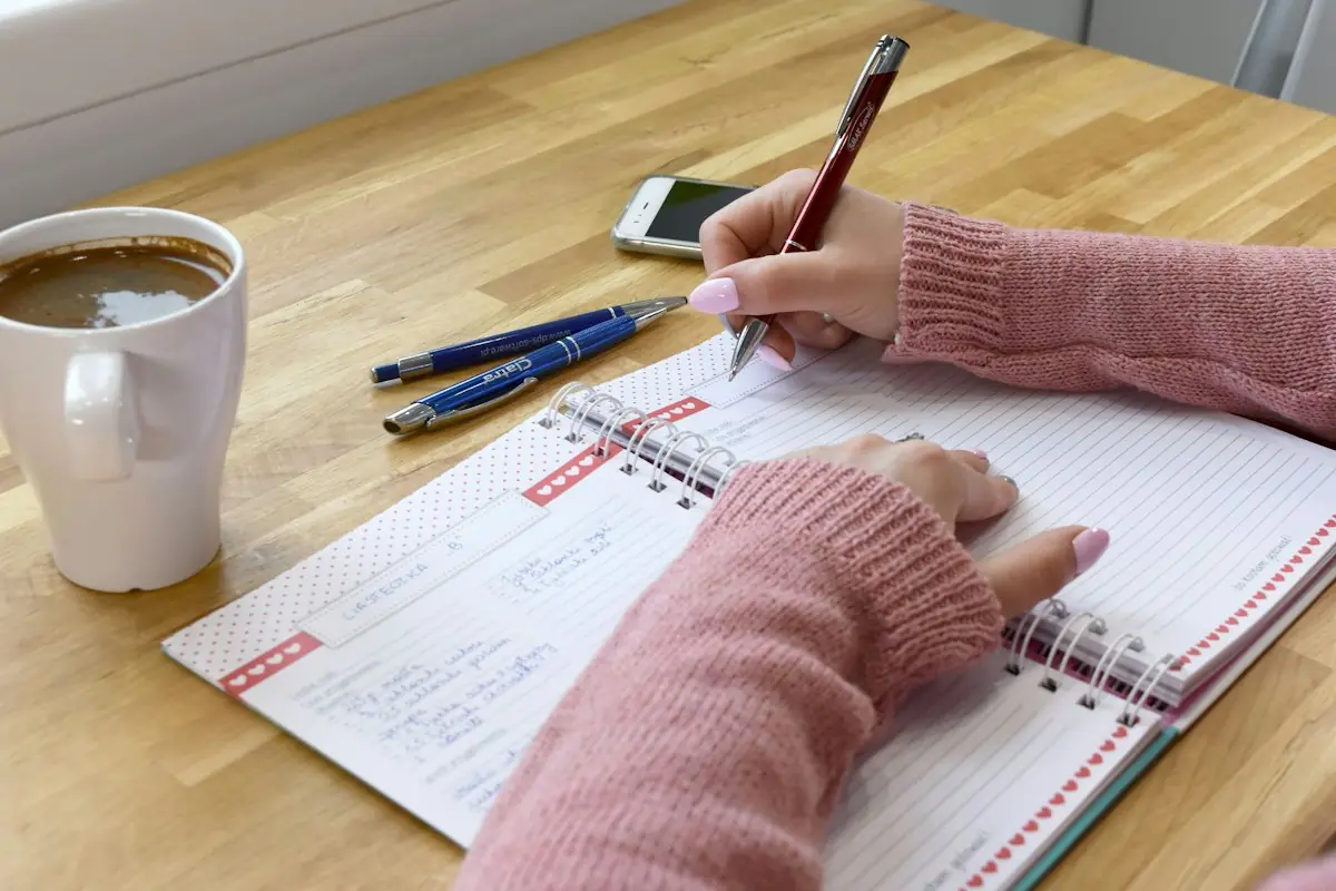 Une main qui recopie une liste de courses dans un carnet, à côté d'une tasse de café
